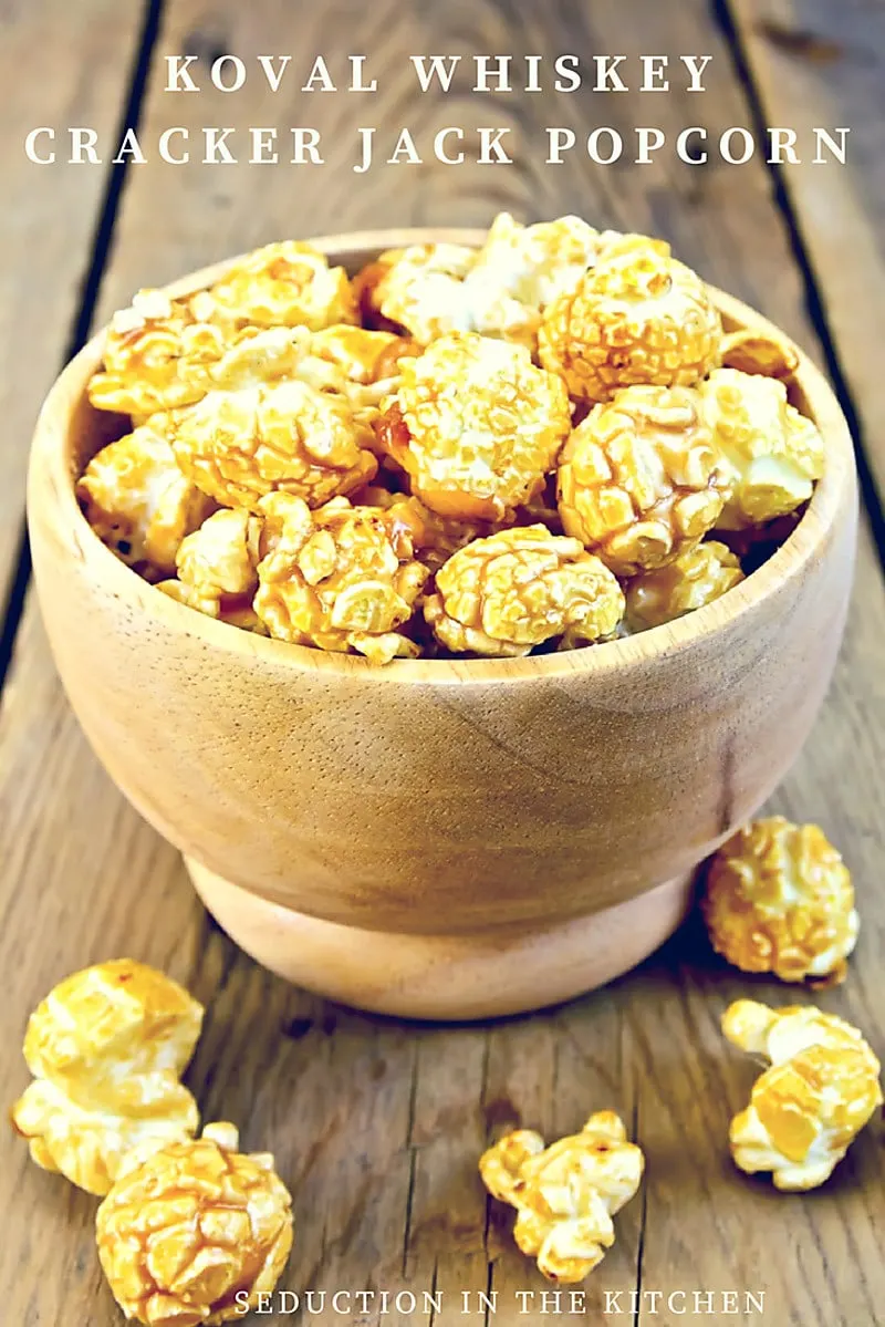 Koval Whiskey Cracker Jack Popcorn is an alcohol-infused caramel update on a copycat Cracker Jack recipe. This caramel glazed popcorn is perfect for a girl's night in. Looking for a fun popcorn snack, look no further than this cracker jack popcorn recipe Koval whiskey cracker jack popcorn in wooden bowl on table with popcorn scattered