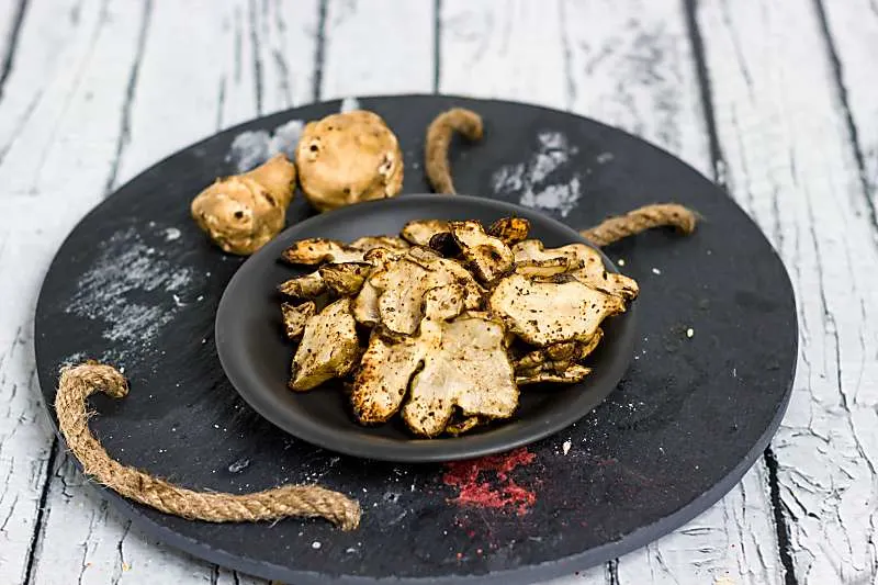 Baked Rosemary And Olive Oil Sunchoke Chips is a healthier alternative to baked potato chips! These nutty flavored baked chips have a great taste with the rosemary salt that flavors this snack Baked Rosemary And Olive Oil Sunchoke Chips long view on table on black plate