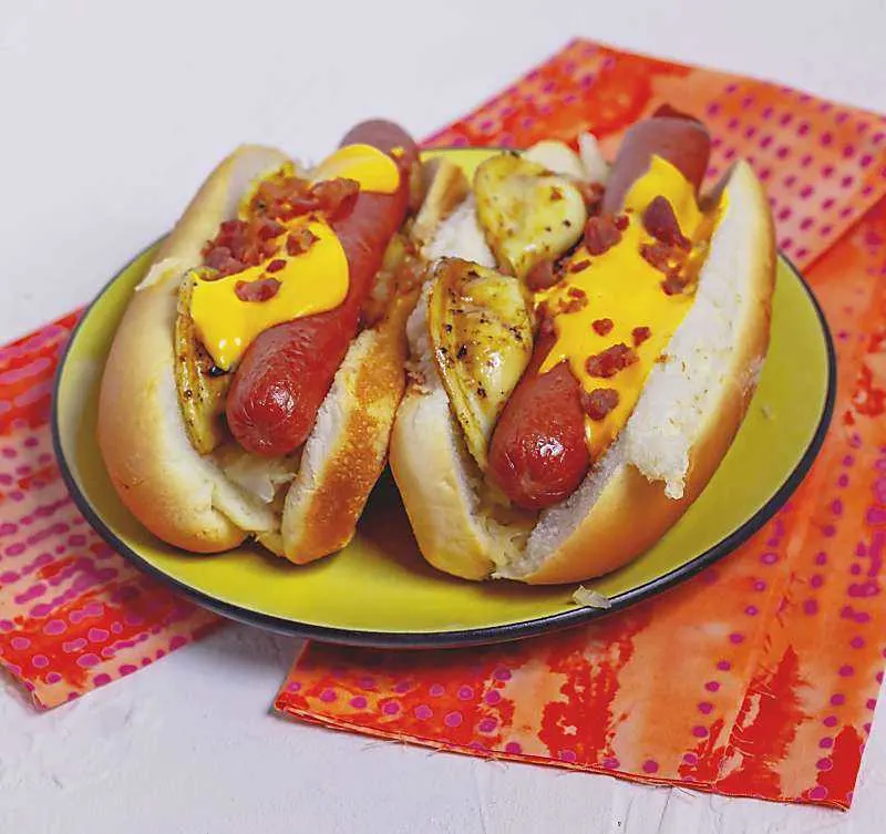 The Pierogi Hot Dog is inspired by ballpark food. This over the top hot dog has sauteed onions, sauerkraut, and of course pierogies! 2 pierogi hot dogs on yellow plate on orange napkin