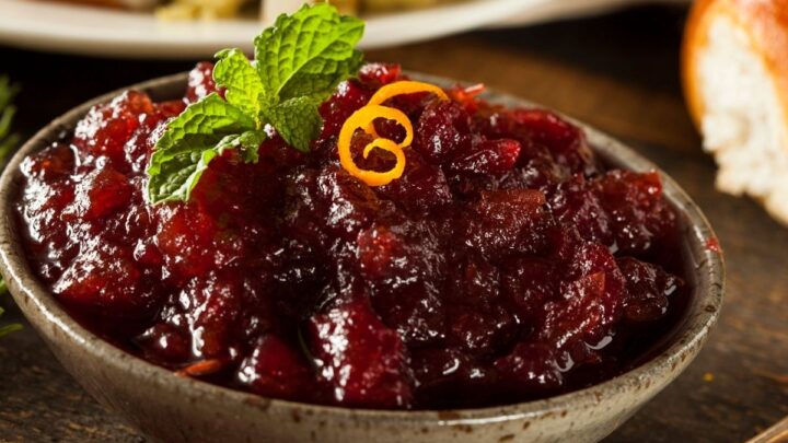 Cranberry sauce in grey bowl with brown specks orange peel garnish on wood table and tarnish spoon