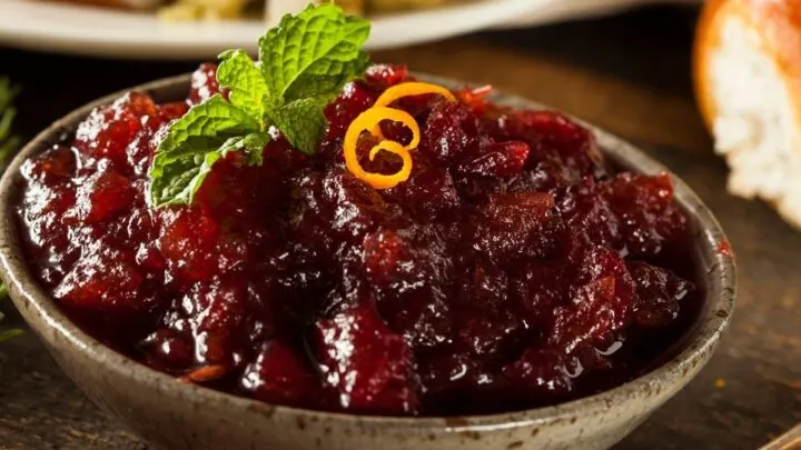 Cranberry sauce in grey bowl with brown specks orange peel garnish on wood table and tarnish spoon