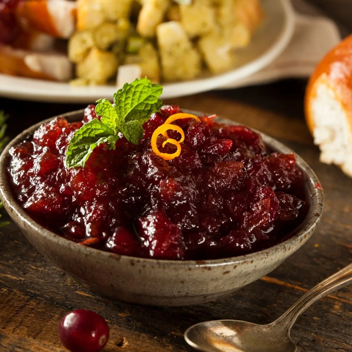 Cranberry sauce in grey bowl with brown specks orange peel garnish on wood table and tarnish spoon