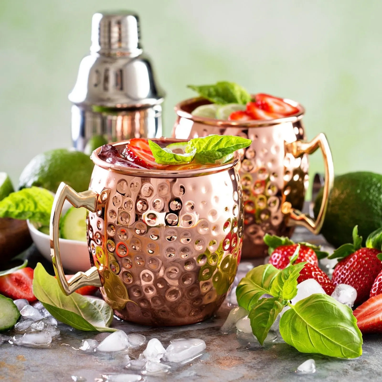 2 strawberry moscow mules with strawberries and mint leaves with cocktail shaker in background